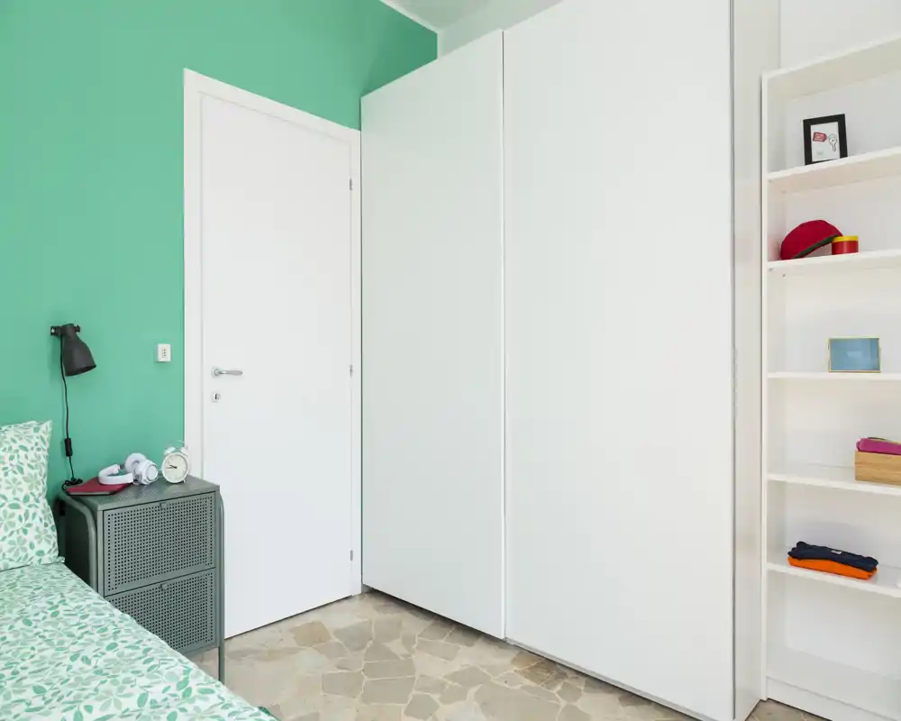 Bright, minimal bedroom corner with a green accent wall, partially visible bed with patterned bedding, bedside table and large white wardrobe.