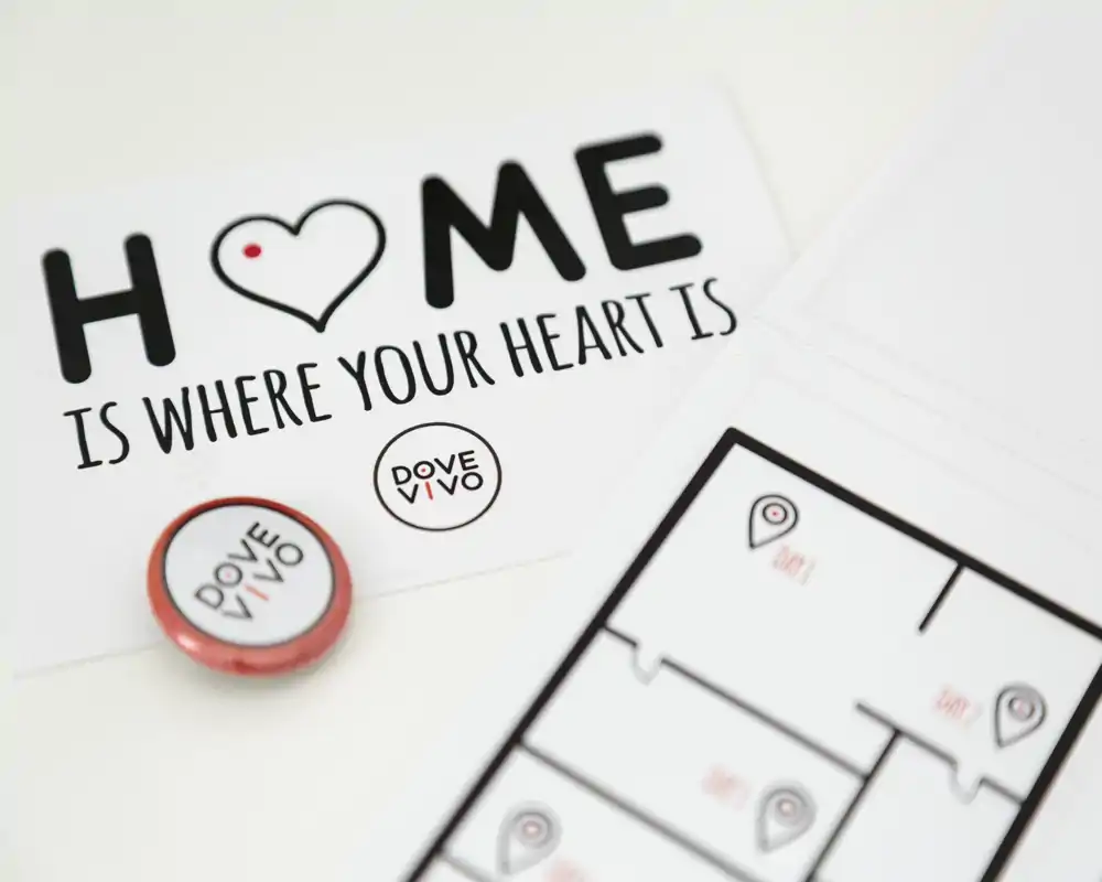 Close-up of printed marketing materials on a white surface: a card reading 'HOME is where your heart is', a round branded badge and a partial floor-plan illustration. This is a product/branding still life, not a room view.