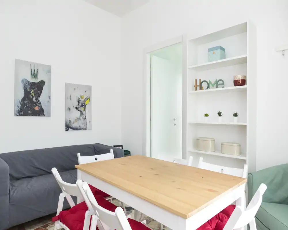 Bright, minimal living/dining area with a wooden table, white chairs and a gray sofa; fresh white walls, shelving and tasteful wall art create a welcoming atmosphere.