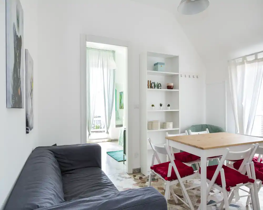 Bright, airy living and dining area with a comfortable sofa, a wooden dining table with red-cushioned chairs, shelving for storage and large windows that flood the room with natural light.