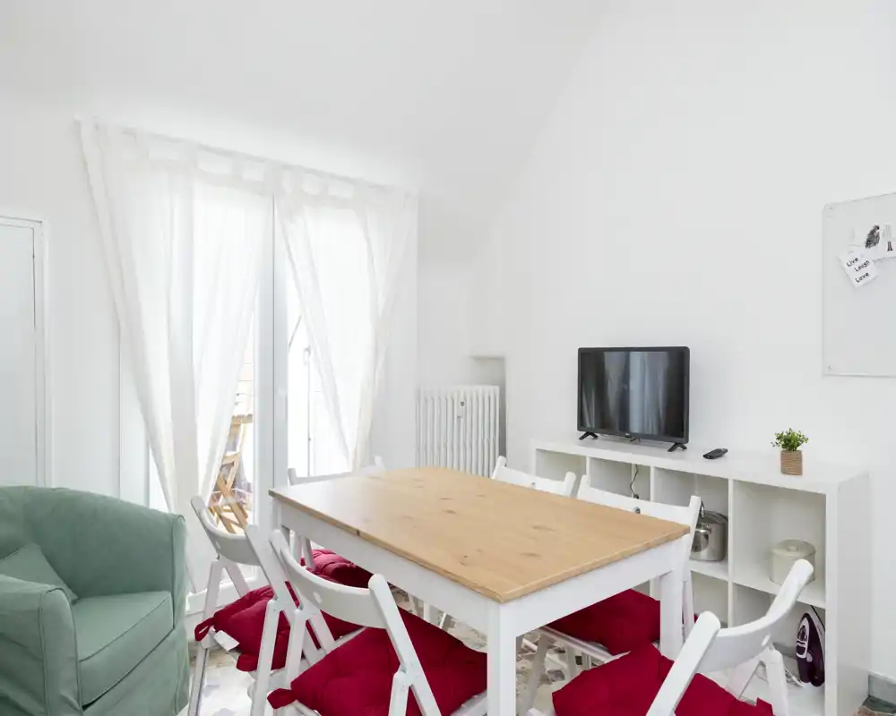 Bright dining area with a wooden table, white folding chairs with red cushions, a TV on a shelving unit and a comfy armchair — ideal for casual meals and small gatherings.