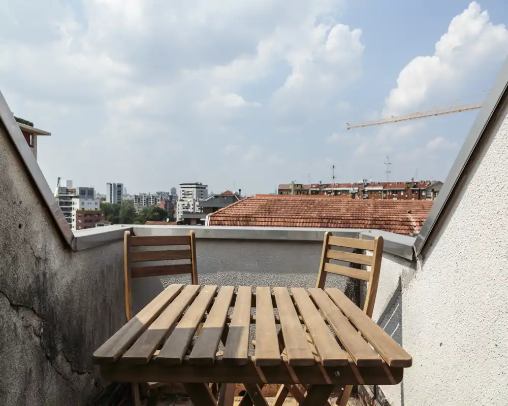 Sunny small terrace with a wooden table and two chairs, overlooking city rooftops and a blue sky with clouds.