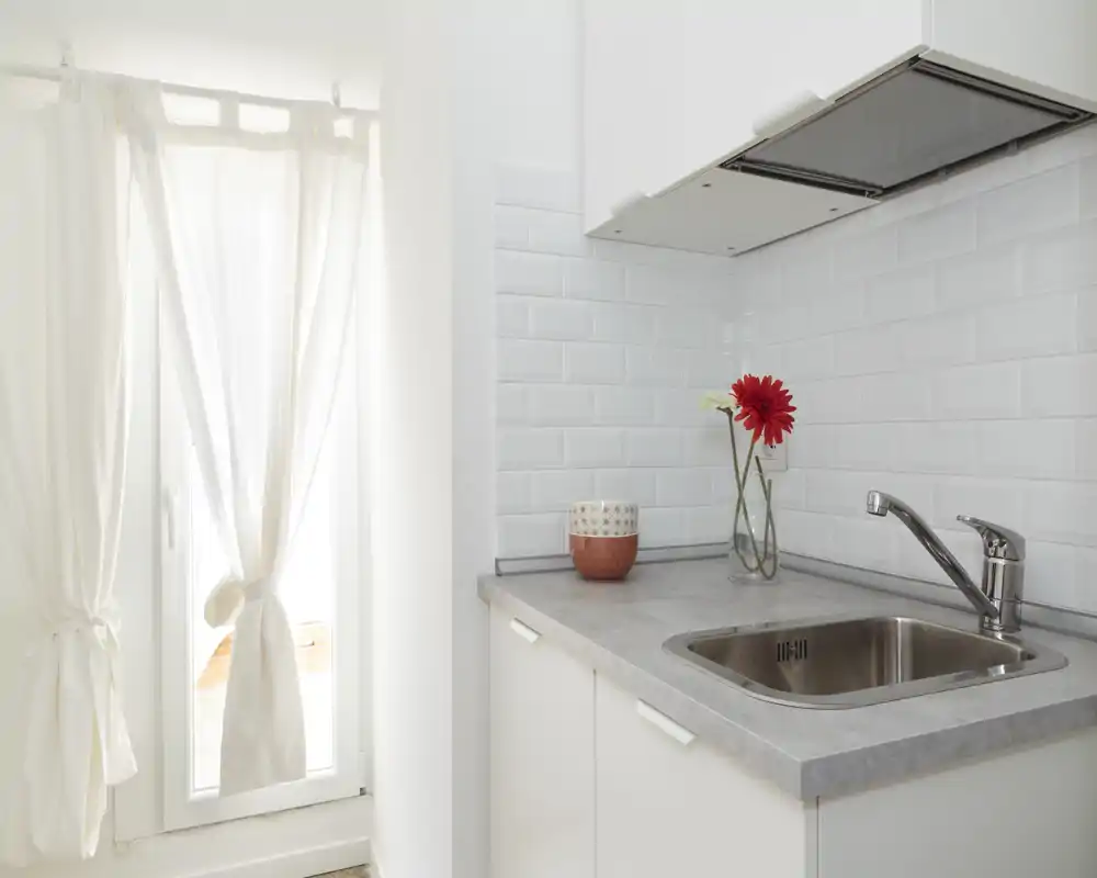 Bright, compact kitchen corner with a stainless-steel sink, light countertop and white subway-tile backsplash; a vase with flowers adds a welcoming touch.