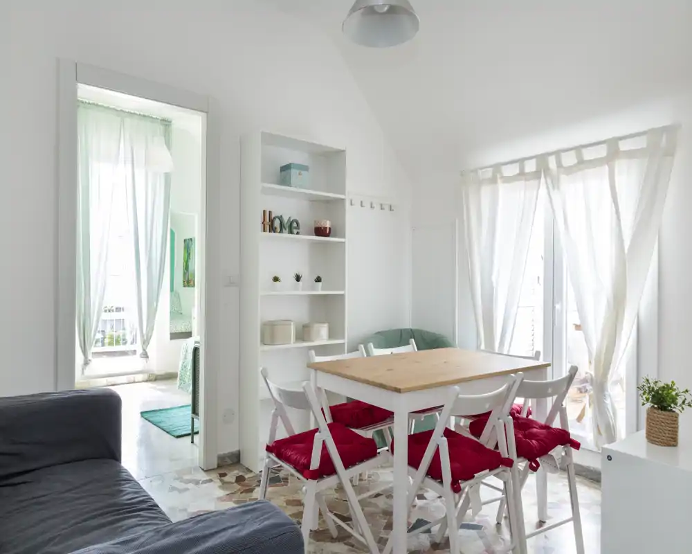 Bright, cozy dining area with a wooden table and white folding chairs dressed with red cushions, set next to large windows letting in abundant natural light.