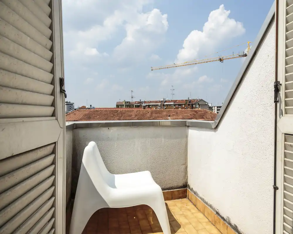 Small private balcony with a single modern white chair, terracotta floor tiles and a view over nearby rooftops under a bright sky.