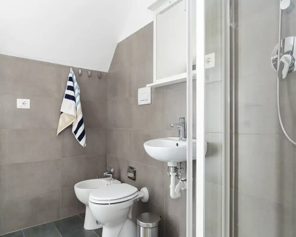 Bright, modern bathroom with wall-mounted sink, toilet, bidet and a glass shower cubicle; neutral grey tiles and tidy fittings.