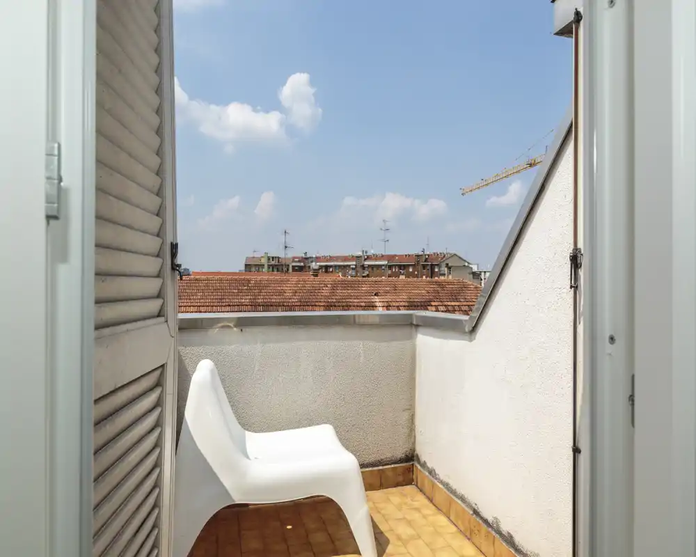 Small private balcony with a white plastic chair, terracotta floor tiles and clear city views under a blue sky — ideal for morning coffee.