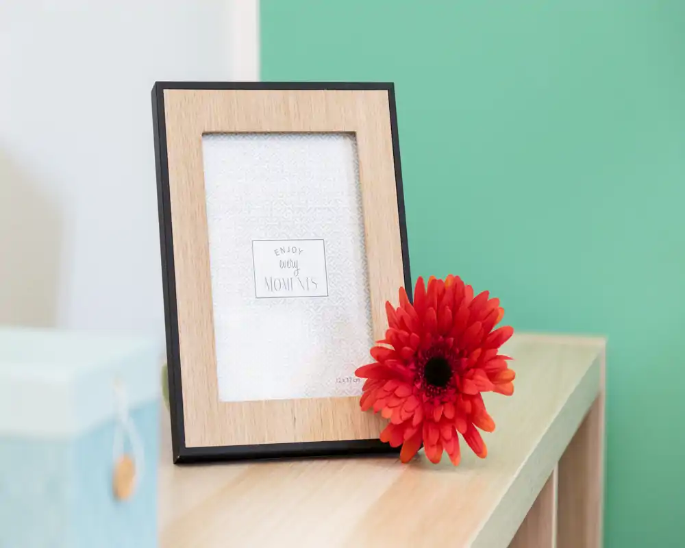 Close-up of a wooden photo frame and a red gerbera on a light-wood shelf against a teal wall. Decorative detail shot suitable for listing accents or staging photos.