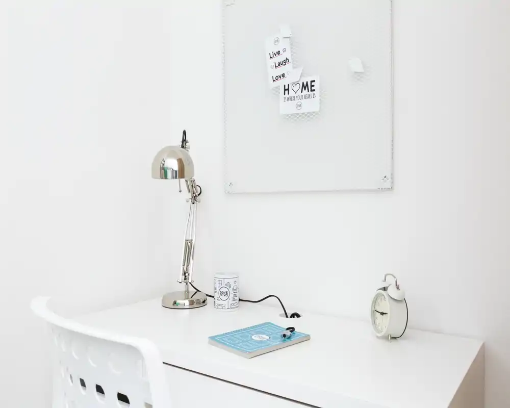 Bright, minimal desk corner with a white chair, metal reading lamp, small notes board and alarm clock — a clean, airy workspace ideal for focused tasks.