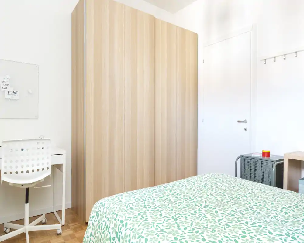 Bright, modern bedroom with a patterned green bedspread, a large light-wood wardrobe, and a compact white desk — clean, airy and well lit.