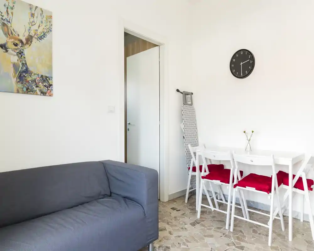 Neat, bright living area with a grey sofa, small dining table with red cushions and minimalist decor — ideal for a compact apartment.