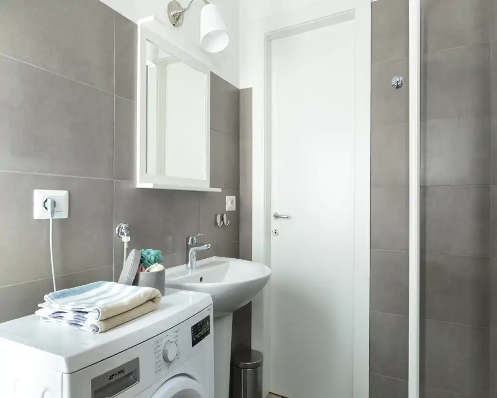 Modern bathroom with gray tiled walls, a white sink and mirrored cabinet, and a washing machine—bright, clean and functional.