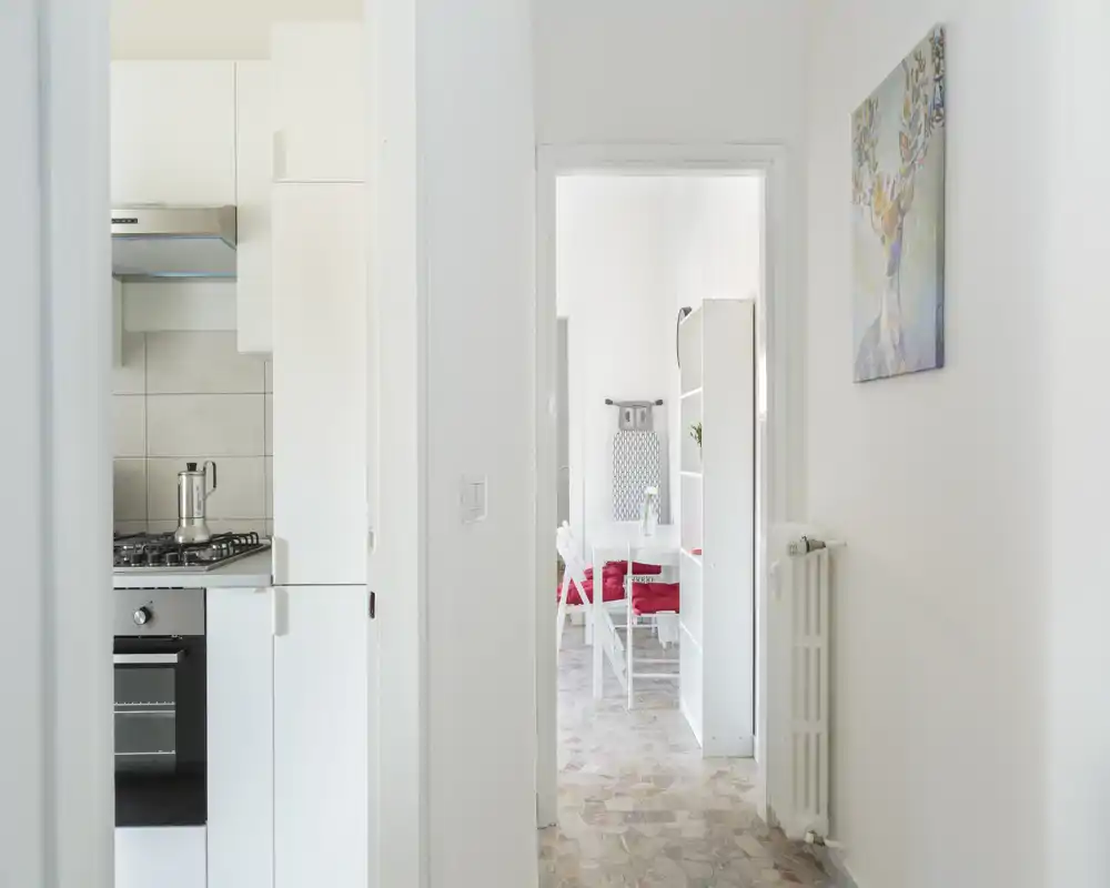 Bright, minimal entrance hall leading to the kitchen and a small dining area; white walls, tiled floor and simple wall art create a clean, welcoming passage.