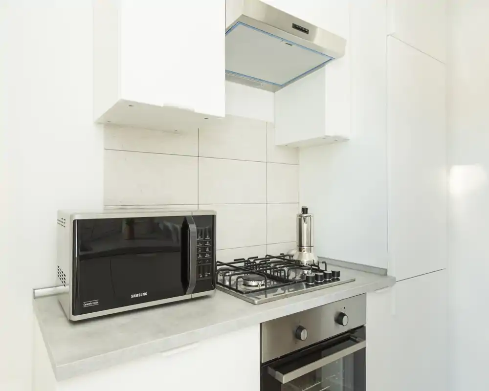 Modern compact kitchen corner with a gas hob, built-in oven, microwave and extractor hood in a bright, minimal design.