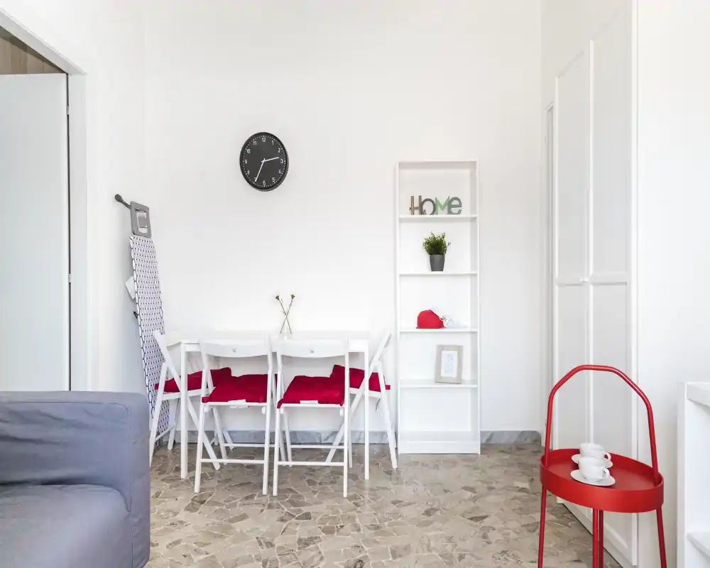 Bright, minimalist dining corner with a small white table, four folding chairs with red cushions, a decorative shelf and a red side table — ideal for compact apartments.