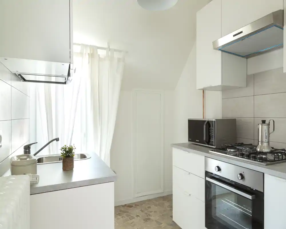 Bright, modern kitchen with white cabinets, gas hob and built-in oven. Natural light from a large window and uncluttered countertops create a fresh, airy space ideal for short-term rentals.