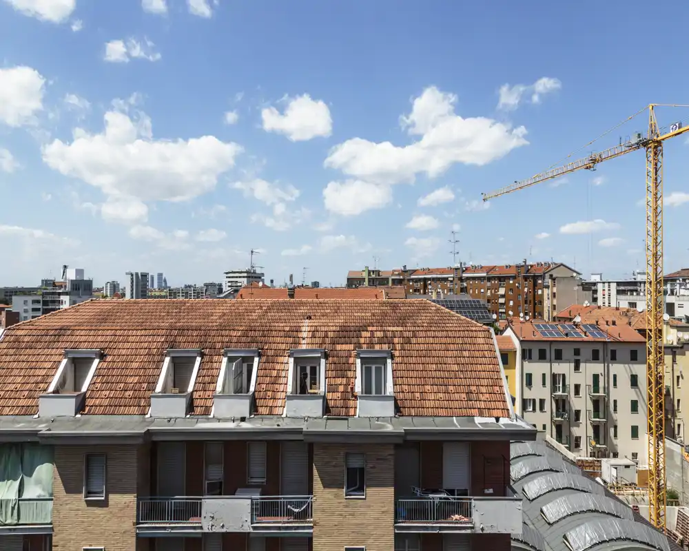 Clear rooftop and city view from a building terrace, showing terracotta roofs, nearby buildings and a construction crane under a blue sky.