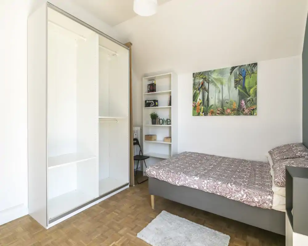 Chambre claire et bien rangée avec lit double, couette à motifs, armoire ouverte et étagères — tons neutres et tableau décoratif apportant une atmosphère chaleureuse.