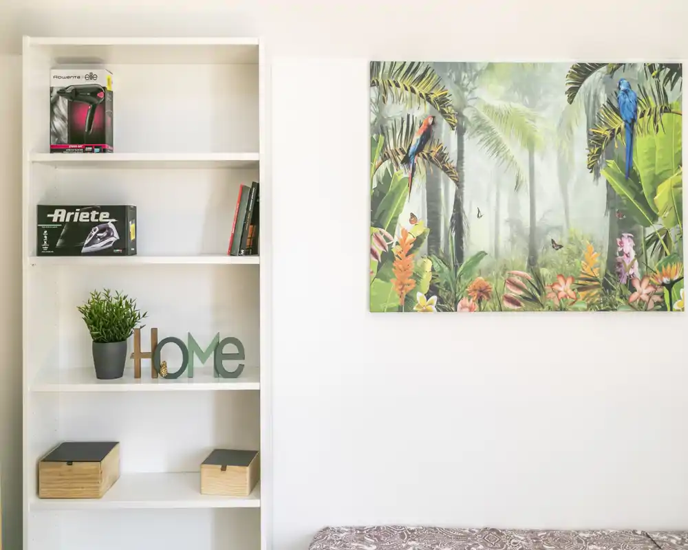 Coin de chambre lumineux et soigné avec une étagère blanche, une plante décorative et une toile tropicale au-dessus du bord du lit. Décor moderne et épuré, l'espace paraît accueillant.
