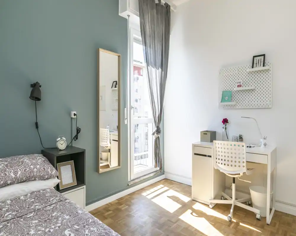 Coin chambre lumineux et moderne avec un lit confortable, une table de chevet et un bureau éclairé par la fenêtre — idéal pour étudier ou se détendre.