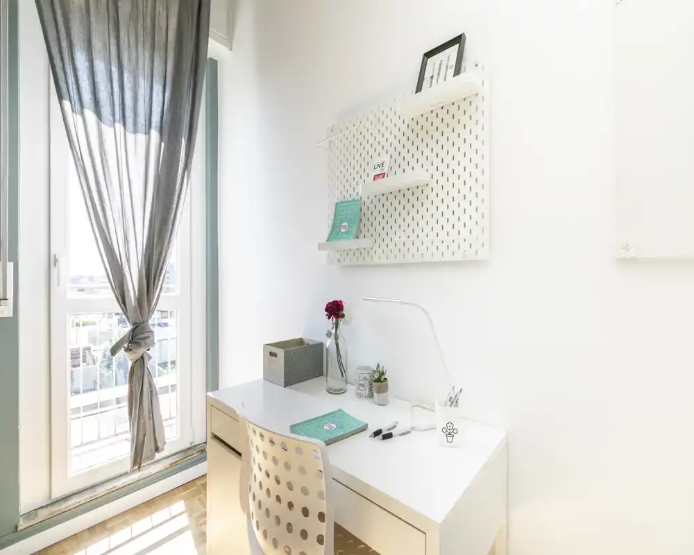 Espace de travail lumineux et minimaliste avec bureau blanc, chaise perforée et lumière naturelle provenant d'une grande fenêtre. Présentation soignée avec lampe de bureau, petites plantes et étagères pour un style moderne et épuré.