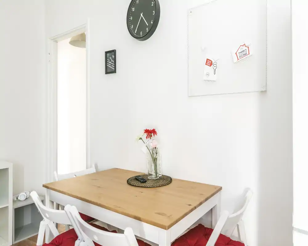 Coin repas lumineux et minimaliste avec table en bois, chaises pliantes blanches et petit vase de fleurs — parfait pour des petits-déjeuners conviviaux.