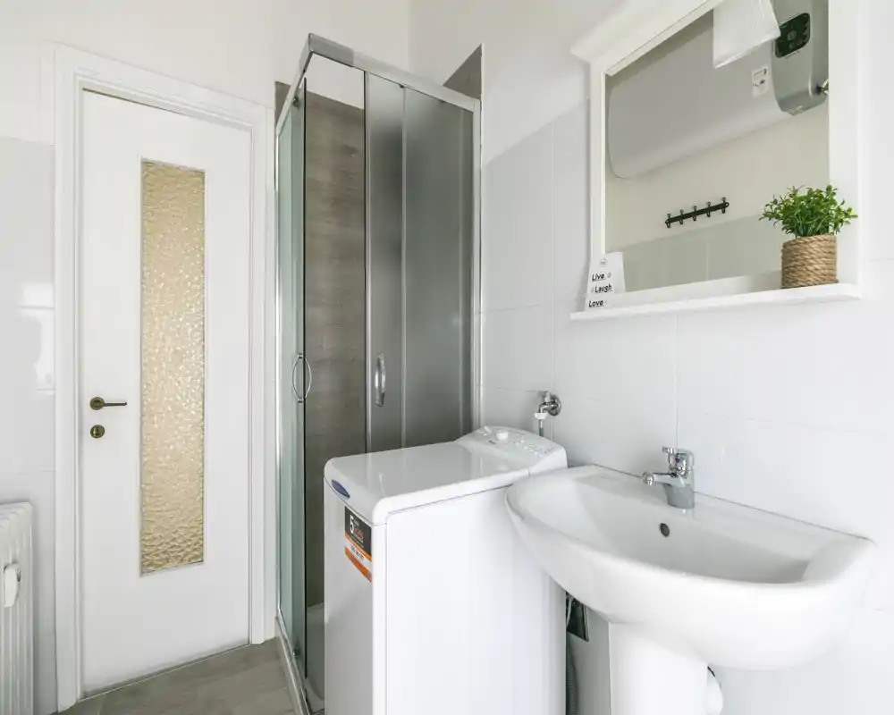 Salle de bains moderne et lumineuse avec cabine de douche en verre, lavabo sur pied et machine à laver; carrelage blanc et armoire miroir pour un espace pratique et soigné.