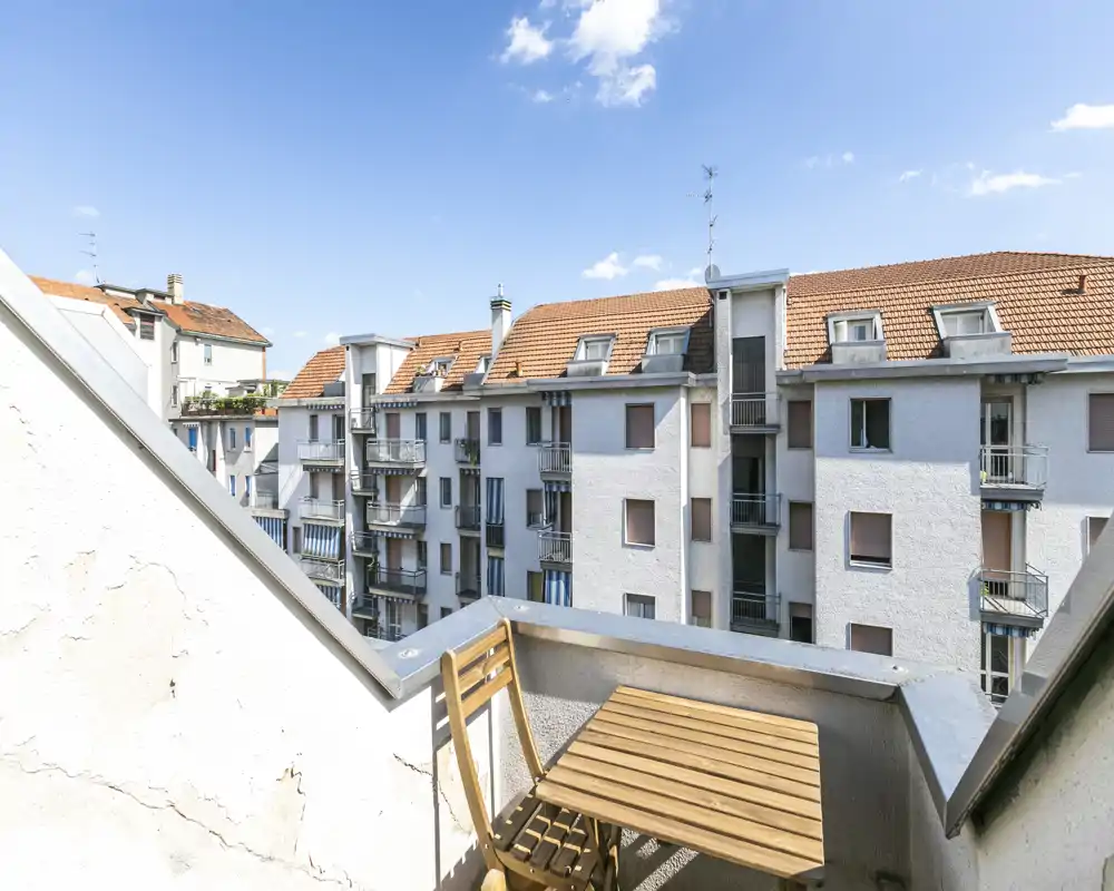 Petit balcon ensoleillé avec une table bistrot et une chaise en bois, offrant une vue sur des immeubles voisins sous un ciel bleu lumineux.
