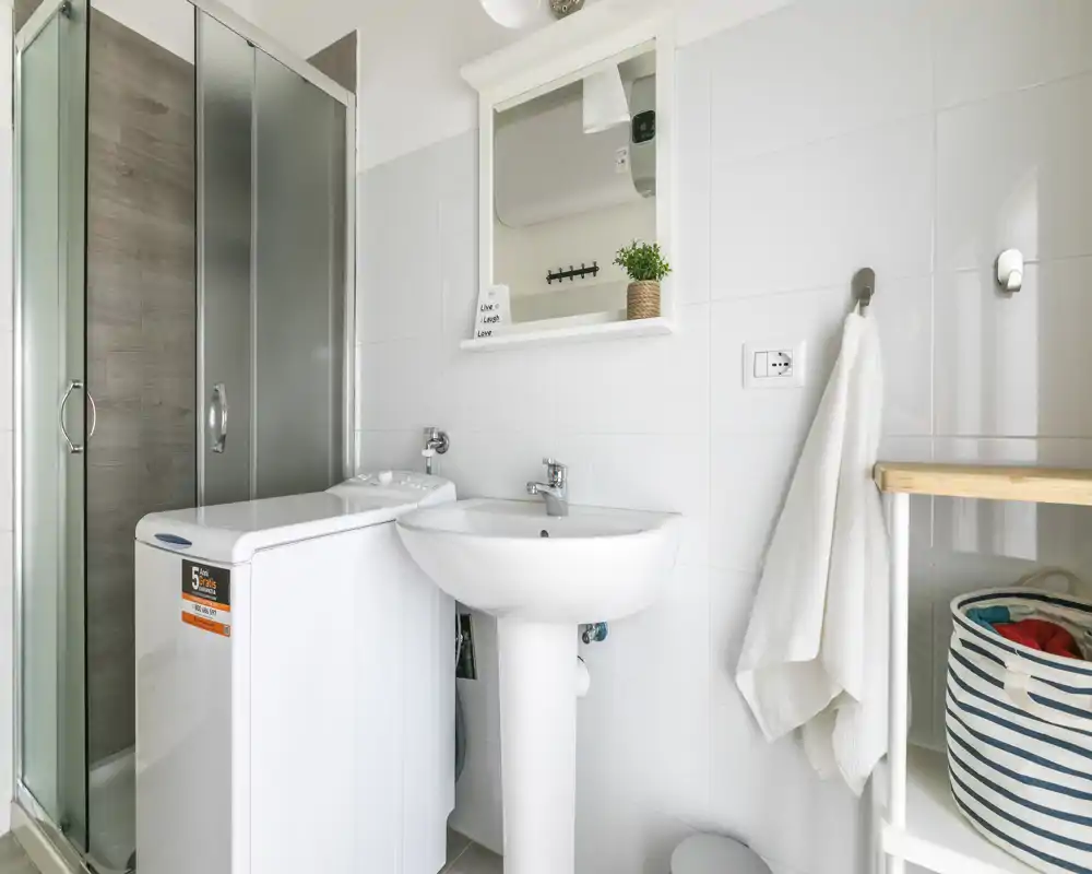 Salle de bain moderne et lumineuse avec lavabo sur pied, armoire-miroir, douche d'angle et une petite machine à laver ; carrelage blanc et rangements simples pour plus de praticité.