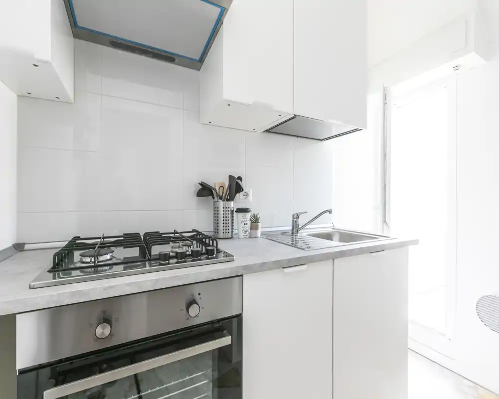 Cuisine moderne et lumineuse avec plaque à gaz, four encastré et évier en inox ; meubles blancs minimalistes et plan de travail clair pour une atmosphère épurée et aérée.