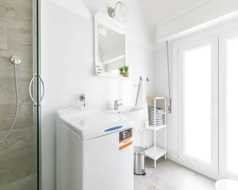Salle de bain moderne et lumineuse avec machine à laver, lavabo, miroir et porte de douche en verre ; carrelage clair et grande fenêtre offrant une atmosphère fraîche et propre.