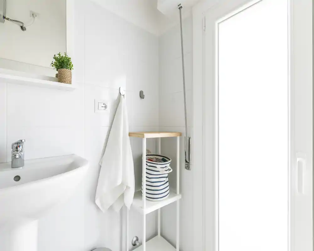 Salle de bains compacte et lumineuse avec lavabo sur colonne, patère pour serviette, étagère et zone de douche à côté d'une porte vitrée ; carrelage blanc et accessoires minimalistes.