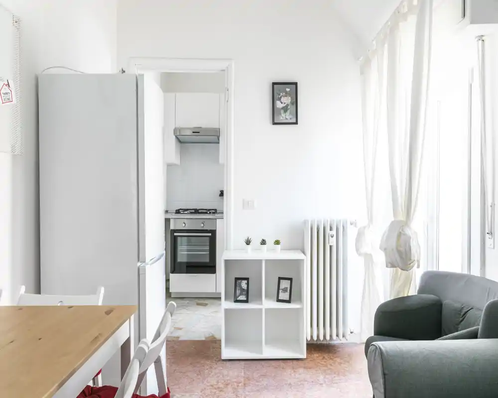 Espace de vie lumineux et minimaliste avec un fauteuil confortable, une petite étagère avec photos encadrées et plantes, et une table à manger partiellement visible; de grandes fenêtres aux rideaux légers apportent une belle lumière naturelle.