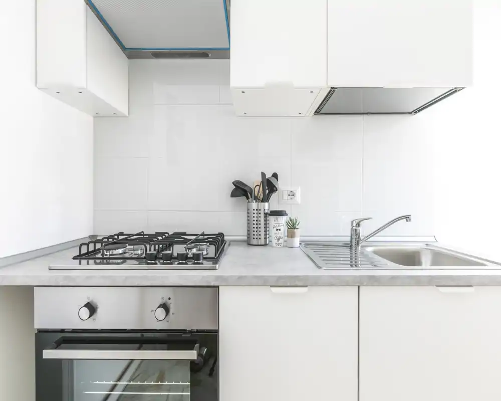 Cuisine moderne et lumineuse avec plaque à gaz, four encastré et évier en inox. Surfaces blanches épurées et style minimaliste avec un porte-ustensiles et une petite plante.