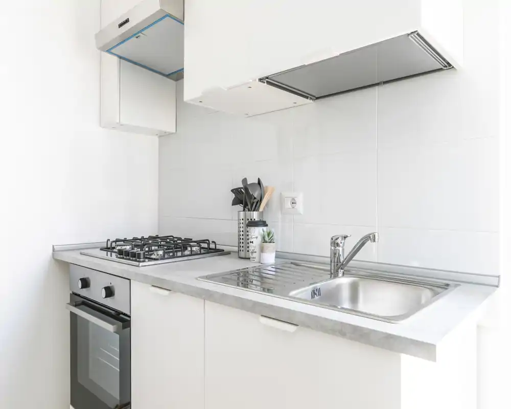 Cuisine moderne et lumineuse avec évier inox, plaque à gaz et four encastré. Rangements blancs minimalistes et petites touches décoratives pour un espace propre et fonctionnel adapté aux locations courte durée.