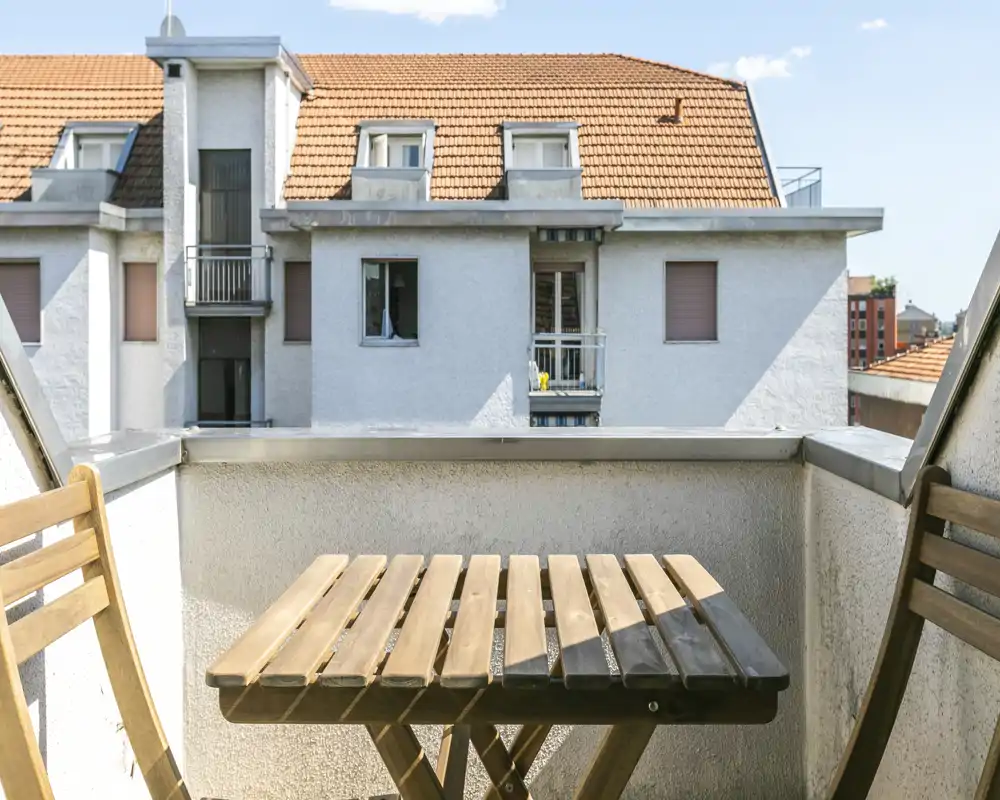 Petit balcon ensoleillé avec une table bistrot en bois et deux chaises pliantes, donnant sur les toits voisins — idéal pour le café du matin.