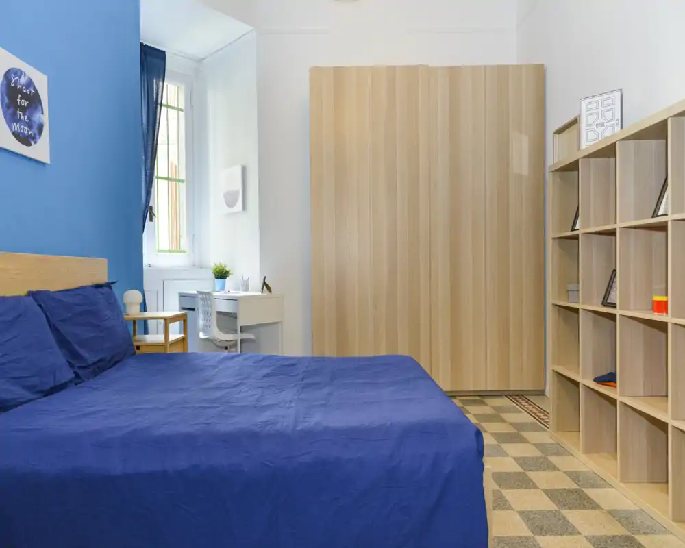 Chambre lumineuse et bien rangée avec un grand lit habillé de bleu, une armoire et des étagères en bois, et un petit bureau près de la fenêtre — idéale pour un séjour confortable.