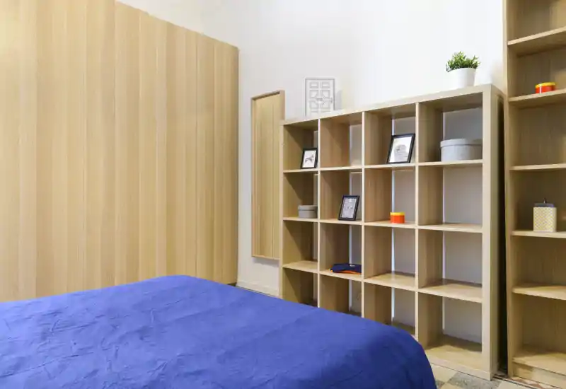 Chambre moderne avec lit recouvert de bleu au premier plan et étagères en bois clair et armoire. Décor lumineux et minimaliste avec rangements en cubes et petits objets décoratifs.