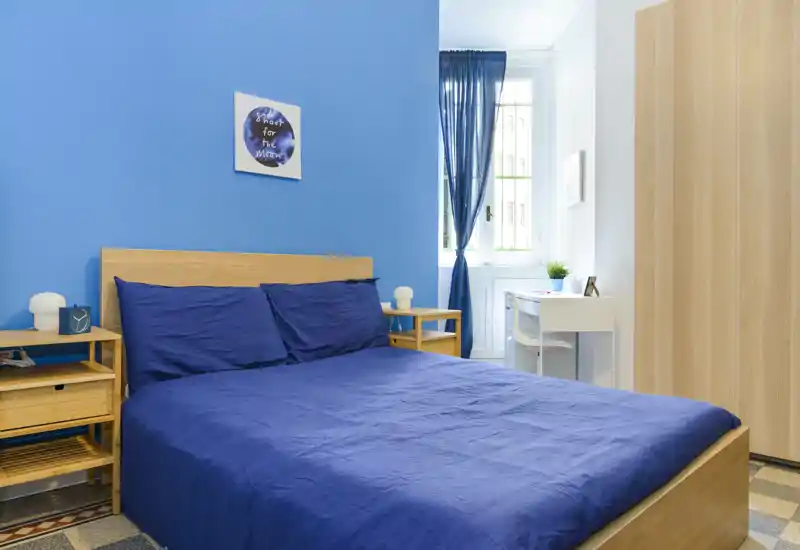 Chambre lumineuse et soignée avec un lit double en bois habillé d'une literie bleu foncé, rideaux assortis et un petit espace bureau près de la fenêtre. La lumière naturelle et le mobilier épuré créent une ambiance moderne et apaisante.