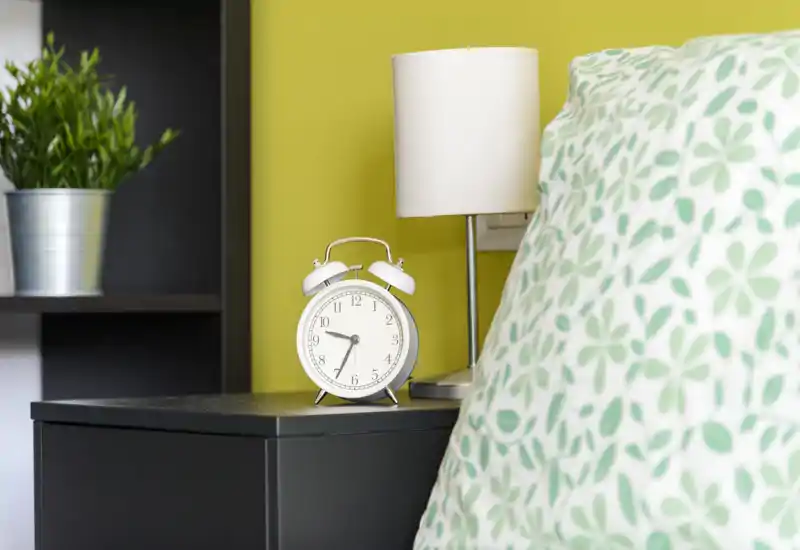 Dettaglio di una camera luminosa e moderna con comodino, sveglia, lampada e cuscino a fantasia: accenti verdi e stile curato, ideale per un annuncio di affitto.