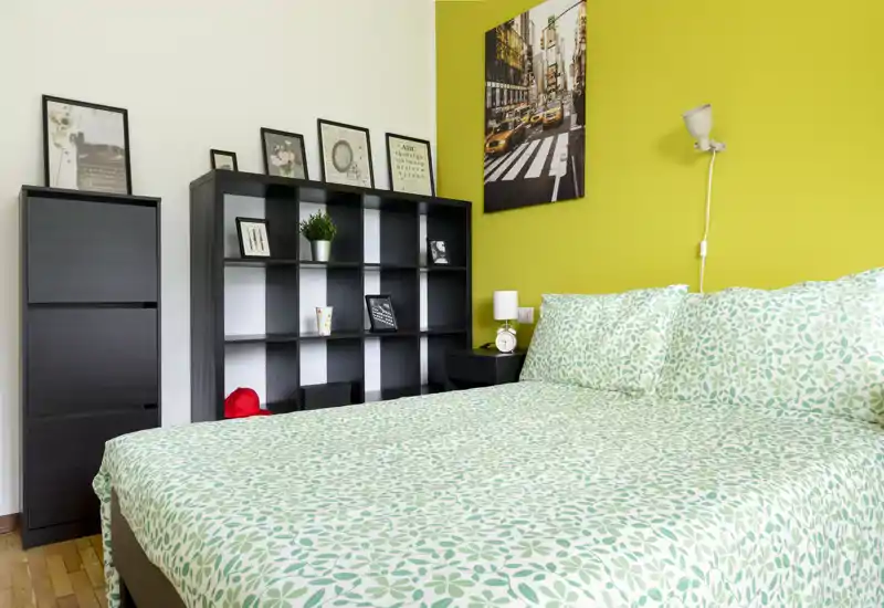 Camera da letto luminosa e ben curata con un ampio letto matrimoniale con copriletto verde a motivo, una libreria nera e comodino su una vivace parete gialla.