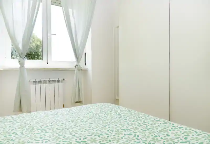 Camera da letto luminosa e minimal con copriletto a motivo in primo piano, tende leggere e una grande finestra che illumina naturalmente l’ambiente. Arredamento pulito e arioso, perfetto per evidenziare luce e semplicità nell’annuncio.