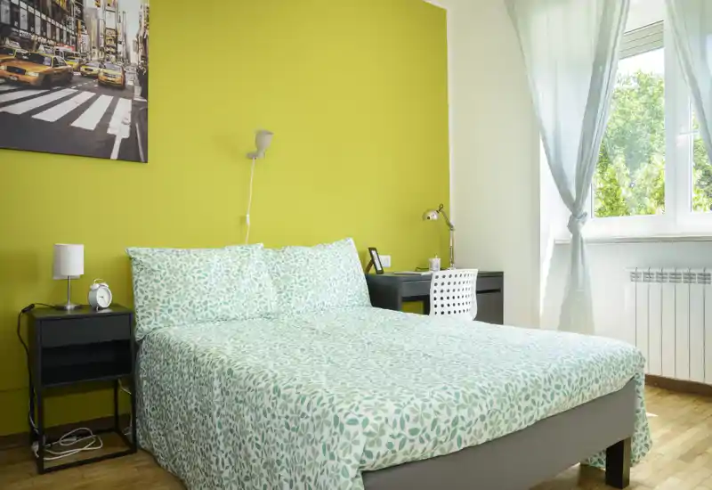 Camera da letto moderna e luminosa con letto matrimoniale con biancheria verde a motivi, comodini, una piccola scrivania e una finestra ampia che introduce luce naturale.