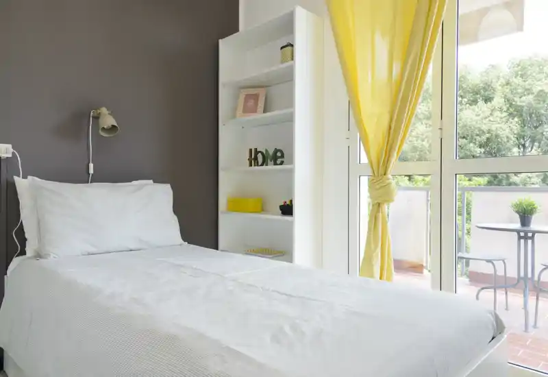 Bright, tidy single bedroom with a white bed, shelving unit and large window opening to a small balcony — ideal for a sunny rental room.