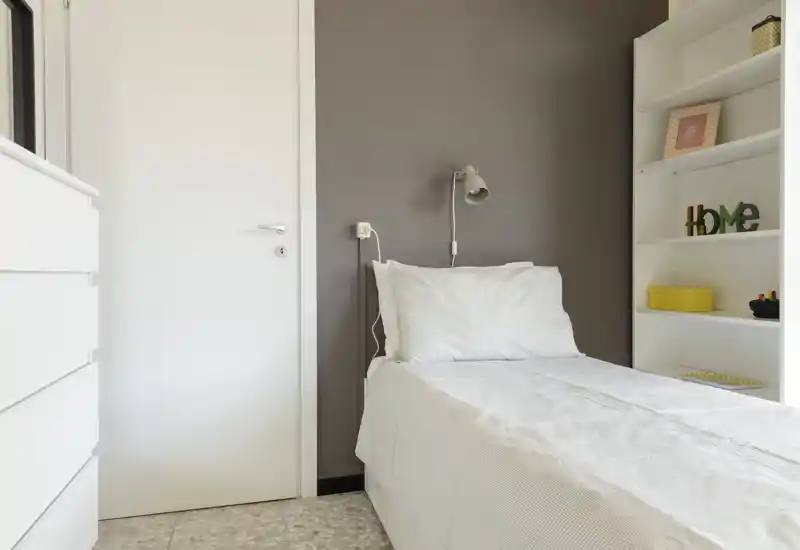 Bright, minimal single bedroom with a neatly made bed against a warm grey accent wall and white shelving for storage.