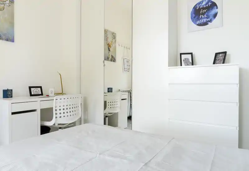 Bright, minimalist bedroom with a neatly made bed, white dresser and a small desk with chair — clean, modern styling ideal for rental photos.