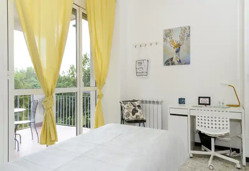 Bright, well-staged bedroom with a neatly made bed, a small desk and chair, warm yellow curtains and a balcony letting in abundant natural light.