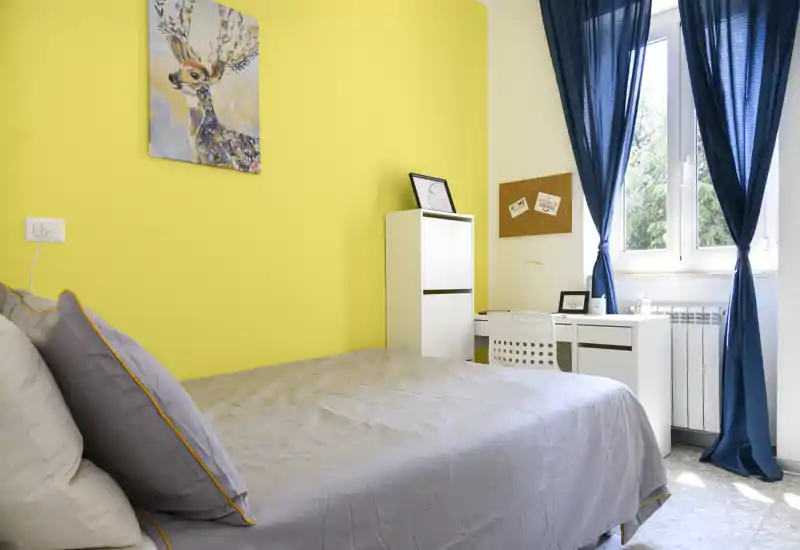 Dormitorio luminoso y bien decorado con cama doble, ropa de cama gris y pared amarilla brillante; escritorio junto a la ventana ideal para trabajar con mucha luz natural.