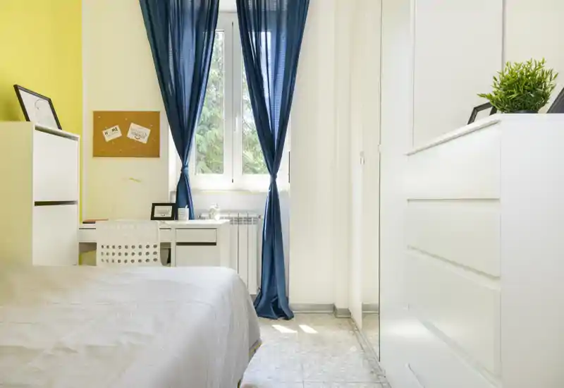 Dormitorio luminoso y ordenado con cama en primer plano, escritorio blanco junto a la ventana con cortinas azules y muebles de almacenaje modernos.