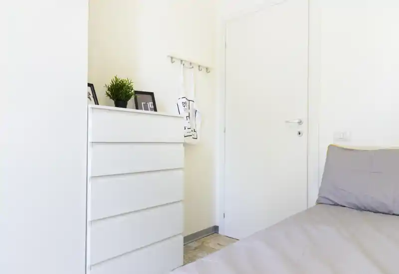 Rincón de dormitorio luminoso y minimalista con la esquina de una cama visible, cómoda blanca y decoración sencilla: limpio, moderno y bien iluminado.